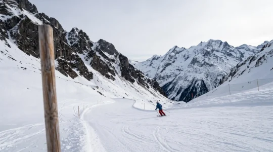 Piste de ski balisee serpentant dans un massif alpin, avec un piquet de signalisation flou au premier plan symbolisant une vigilance necessaire malgre le balisage